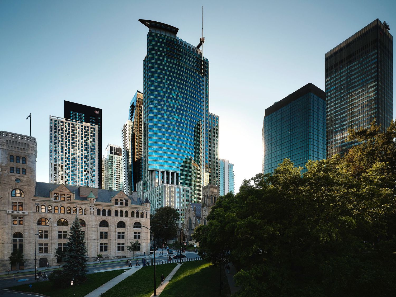 Modern glass skyscrapers and historic stone buildings line a green urban park in downtown Minneapolis under a clear blue sky.