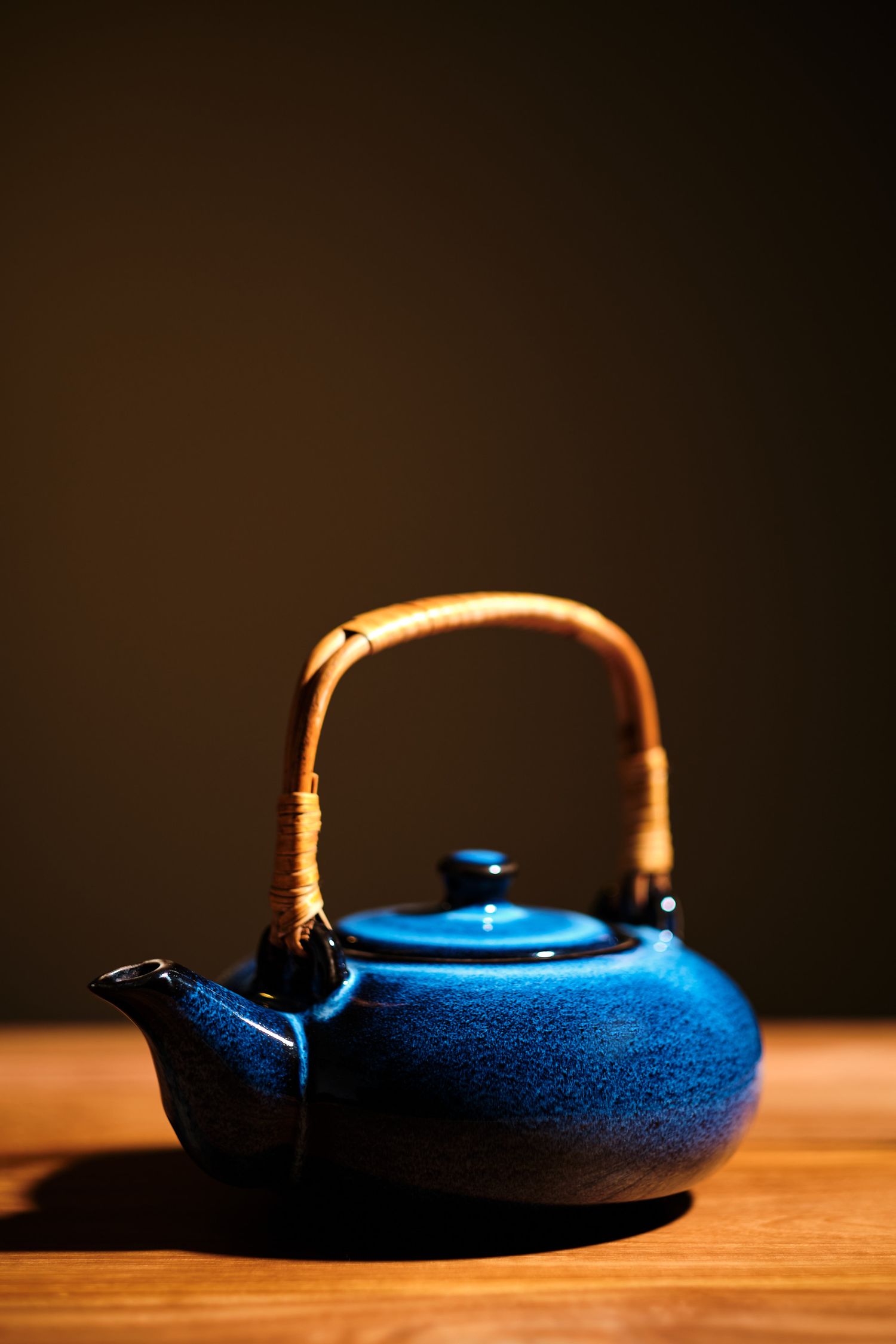 Deep blue teapot with curved spout photographed in low-key lighting on wooden surface.