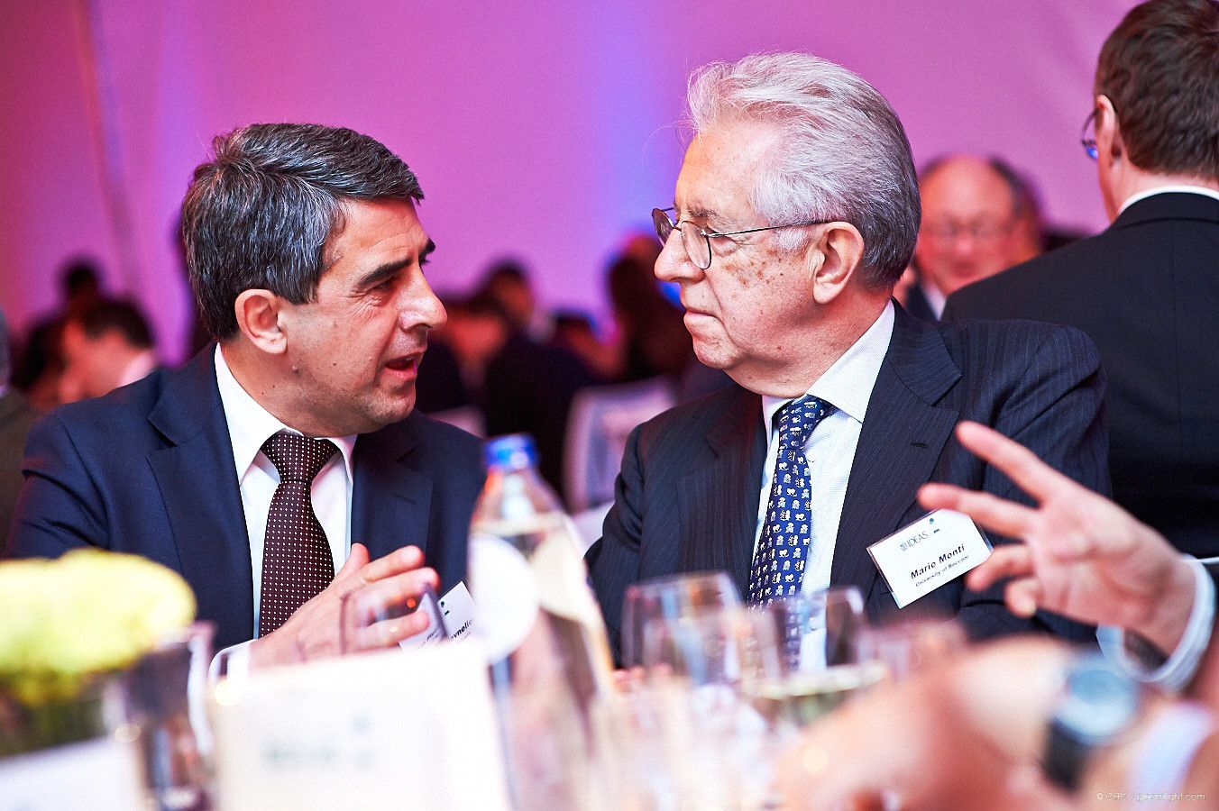 Two people engaged in conversation at an elegant dinner event with pink lighting.