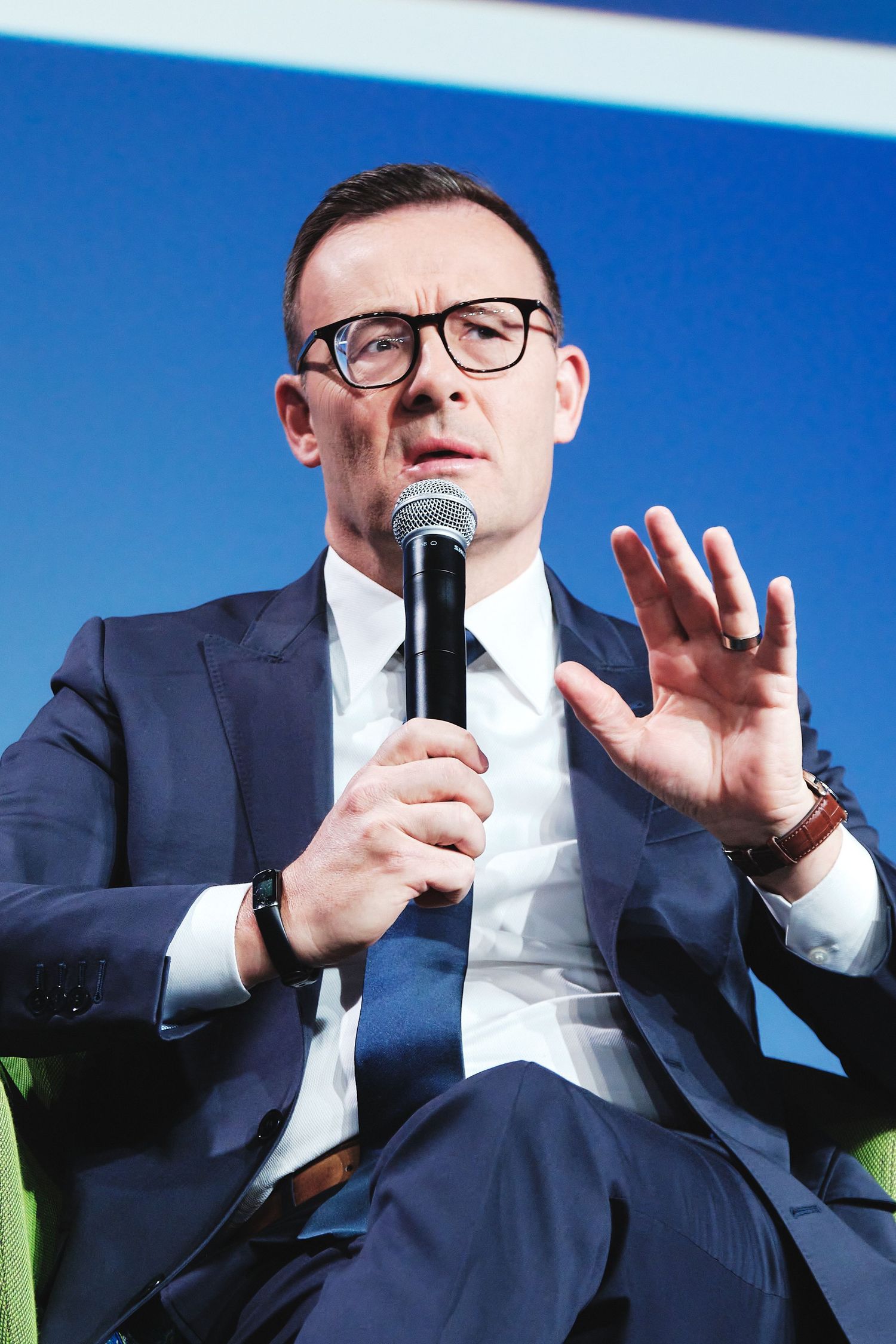 Speaker in a navy suit and glasses gestures while holding microphone during presentation.