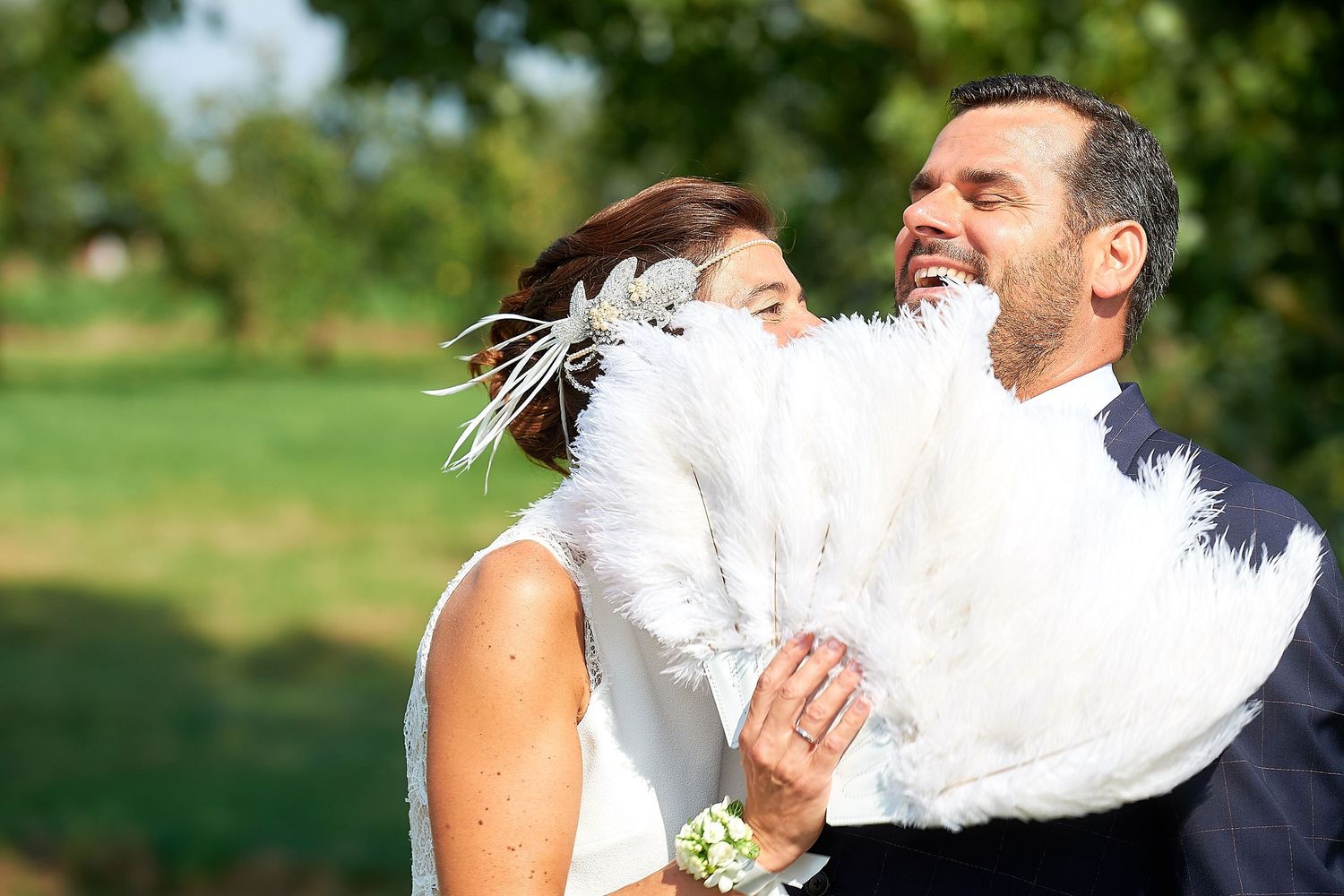 Bride and groom laughing together outdoors, bride holding a white feather fan on a sunny wedding day.