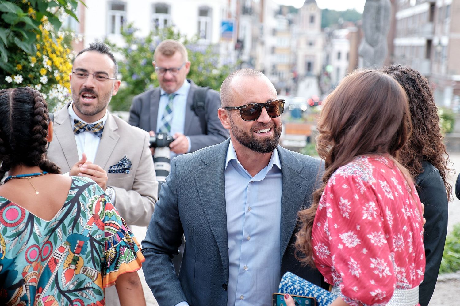 Guests socializing outdoors at a wedding reception, men in suits and bow ties mingling with women in floral dresses.