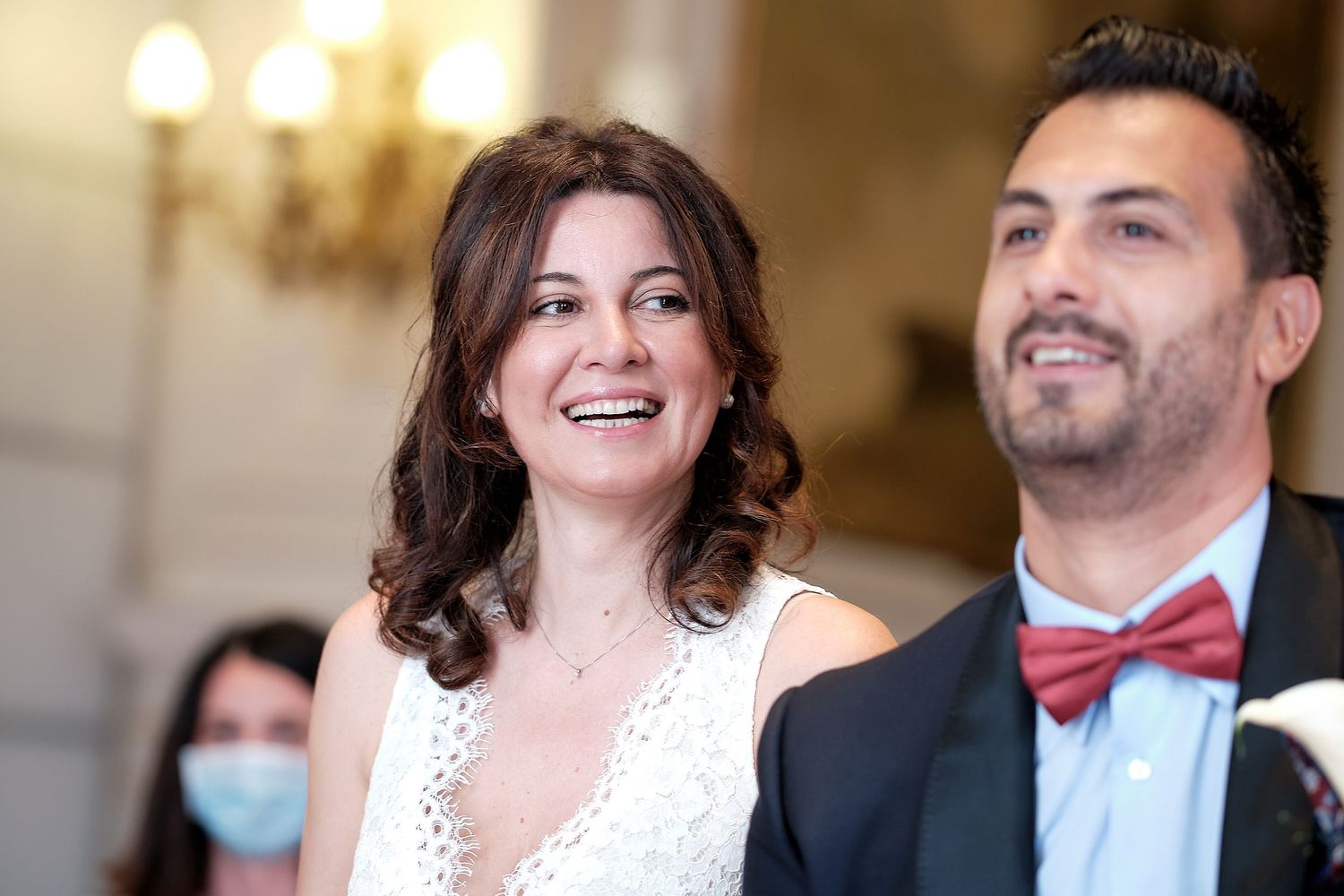Happy couple smiling at a wedding ceremony, woman in white dress, man in red bow tie, indoor venue.
