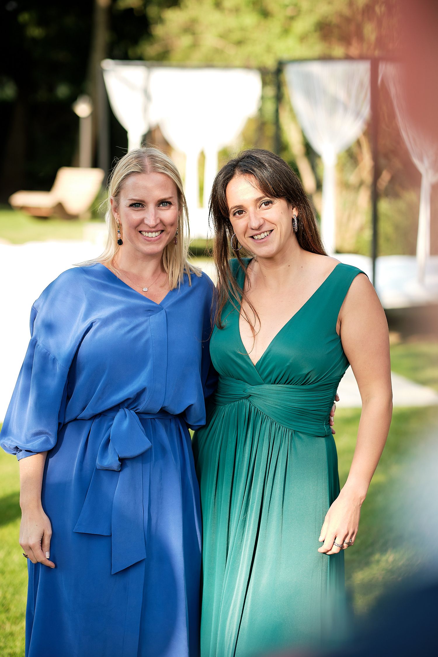 Two women in elegant blue and green gowns smiling outdoors at a formal event.
