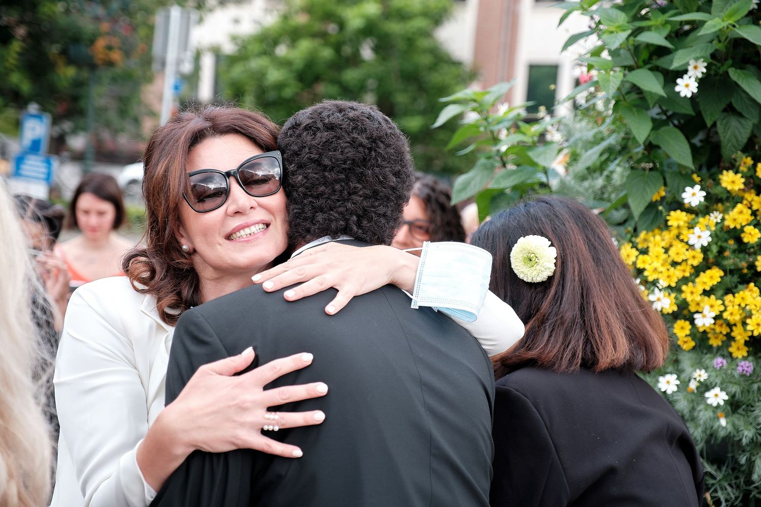 Woman in sunglasses smiling while hugging someone outdoors at a social gathering with flowers in background.