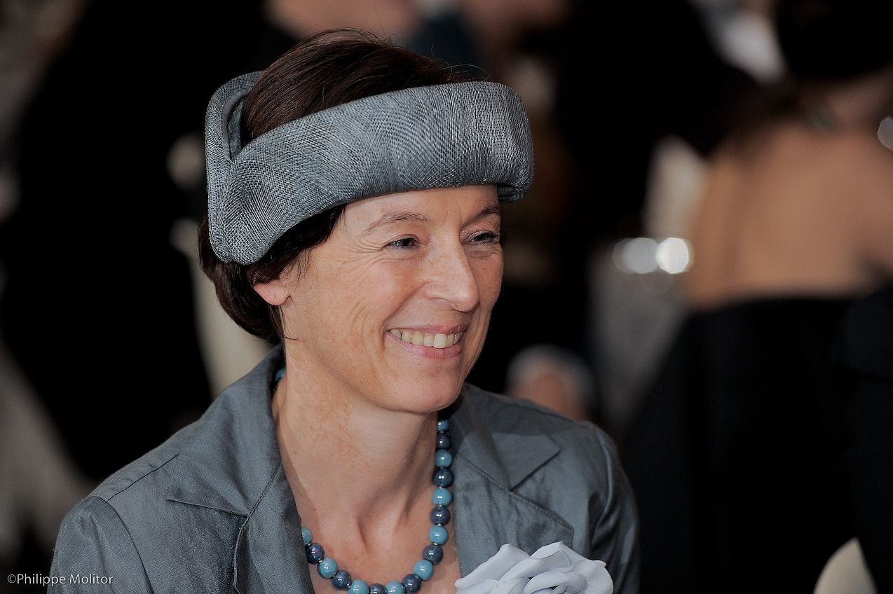 Smiling woman wearing a stylish gray fascinator hat and blue beaded necklace at a formal event.