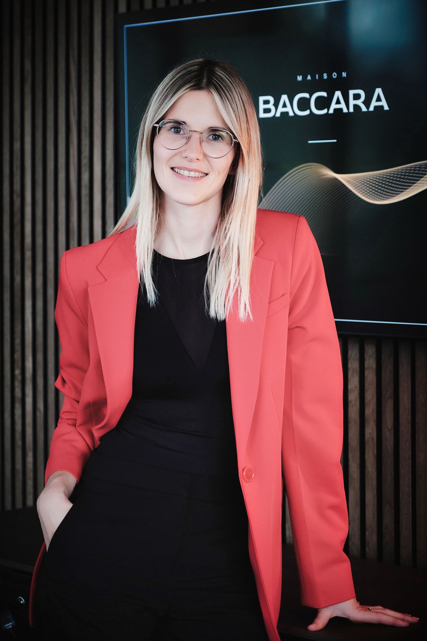 Professional portrait in coral blazer and black dress standing by Baccara company logo.