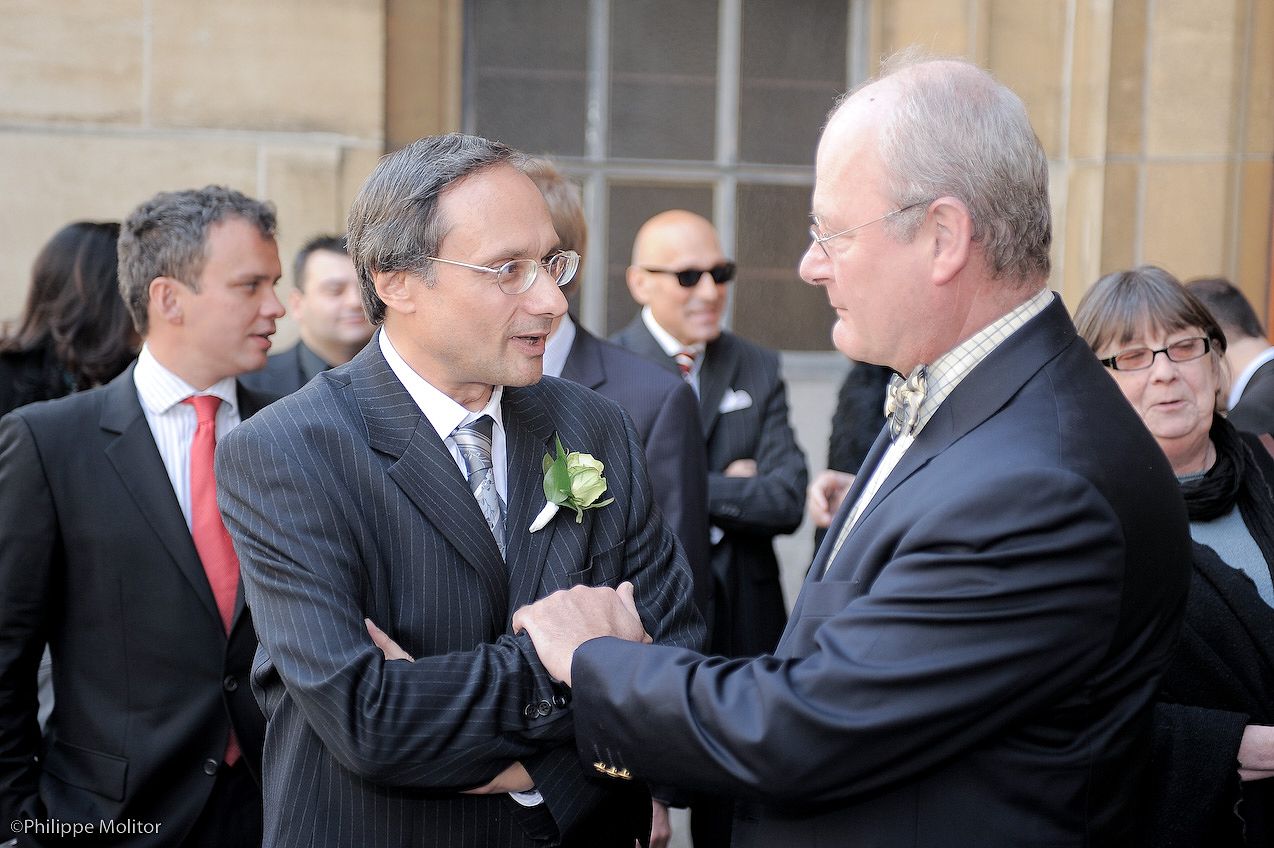 Two men in formal suits shake hands at an outdoor event, with guests gathered in the background.
