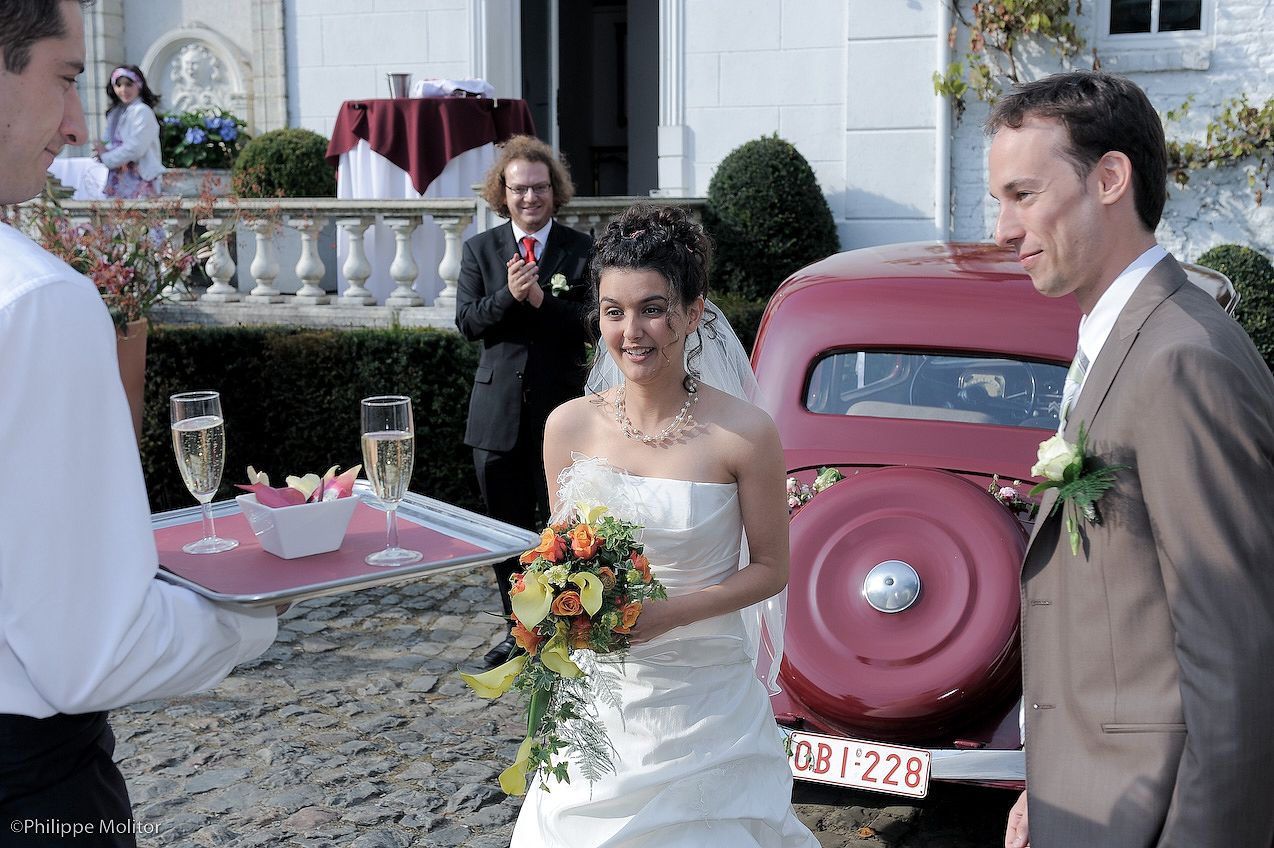 Bride and groom receiving champagne glasses from a waiter near a vintage red car at an outdoor wedding celebration.