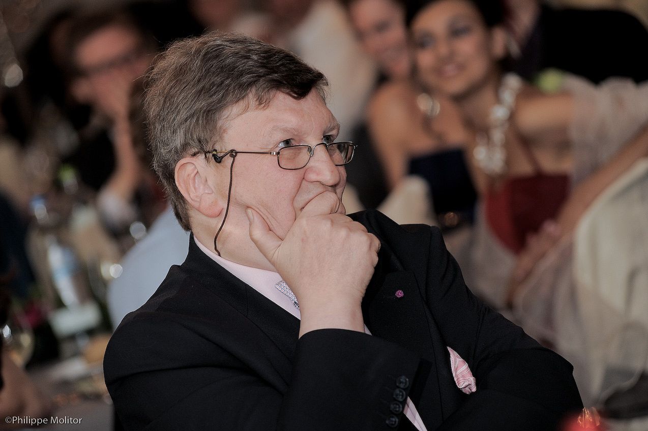 Thoughtful elderly man in glasses and black jacket sitting at a formal event dinner, resting chin on hand.