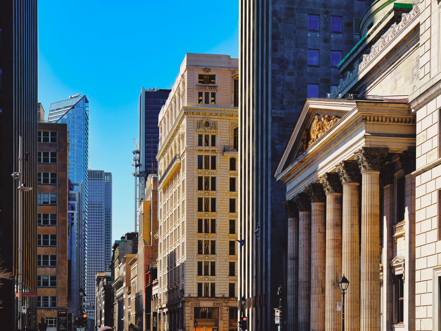 Historic neoclassical buildings with columns line a street in downtown Manhattan with modern skyscrapers in the background.