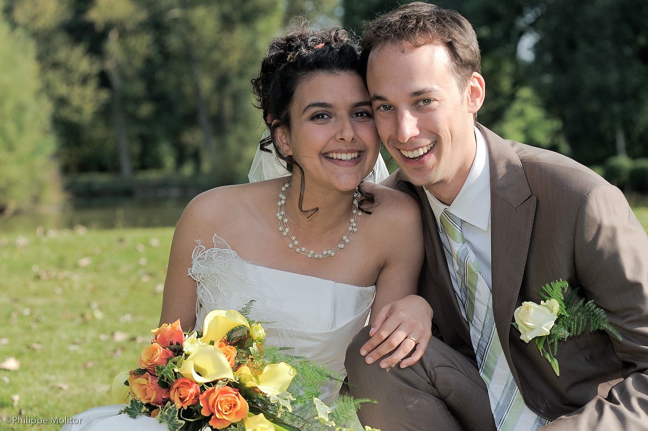 Happy newlywed couple smiling outdoors, bride in white dress holding colorful bouquet, groom in gray suit with boutonniere.