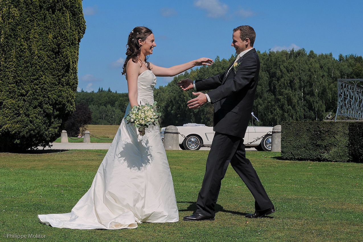 Bride and groom dancing outdoors on green lawn with classic white car and trees in background on sunny wedding day.