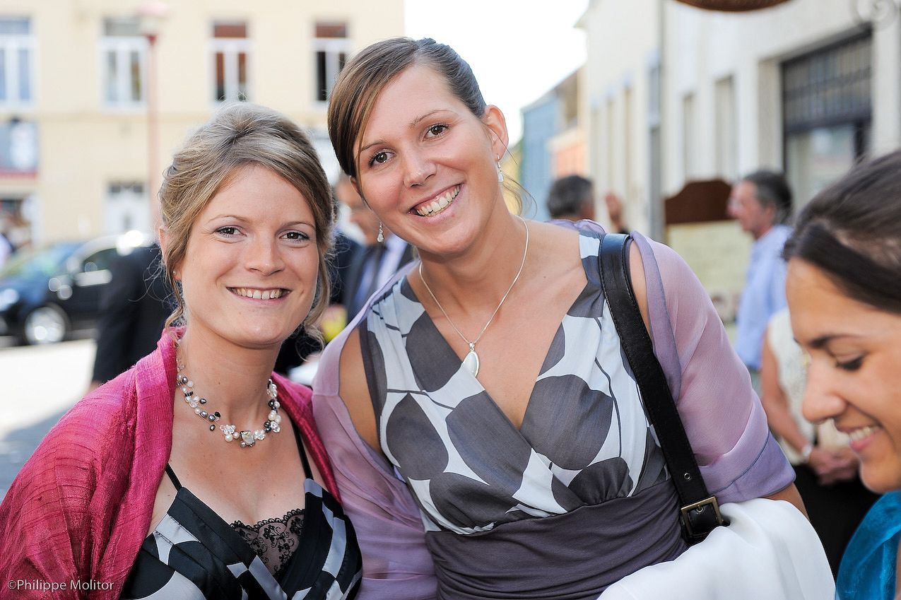 Two smiling women pose together outdoors at a wedding reception on a sunny day.