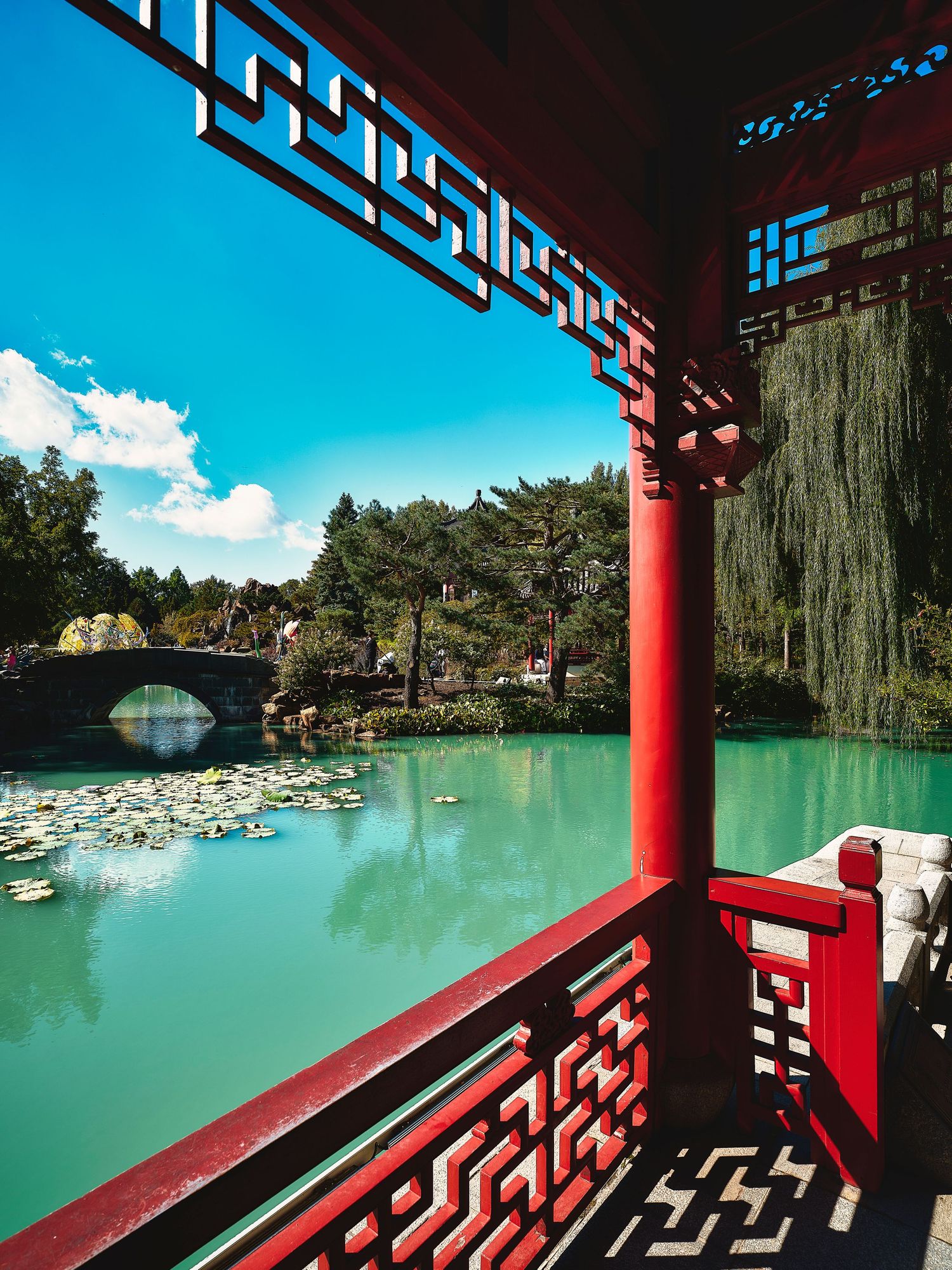 Traditional Asian pavilion with red pillars overlooks a turquoise lake surrounded by trees.