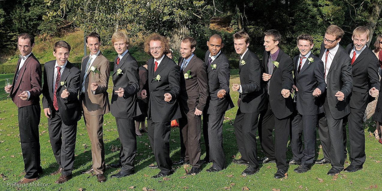 A group of groomsmen in dark suits with boutonnieres posing playfully outdoors at a wedding.