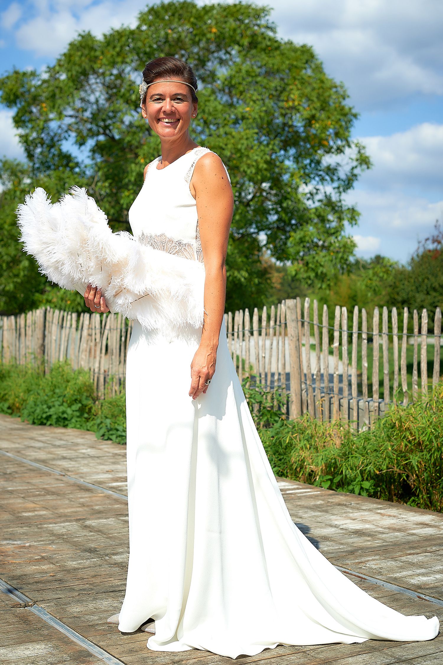 Bride in elegant white gown holding feather bouquet, posing outdoors near wooden fence and green trees on sunny day.