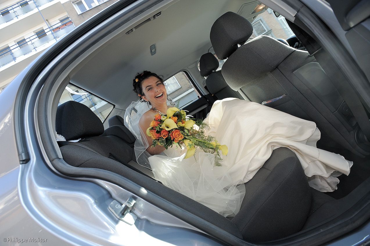 Bride in white wedding dress sitting in car backseat holding colorful bouquet, smiling on wedding day.