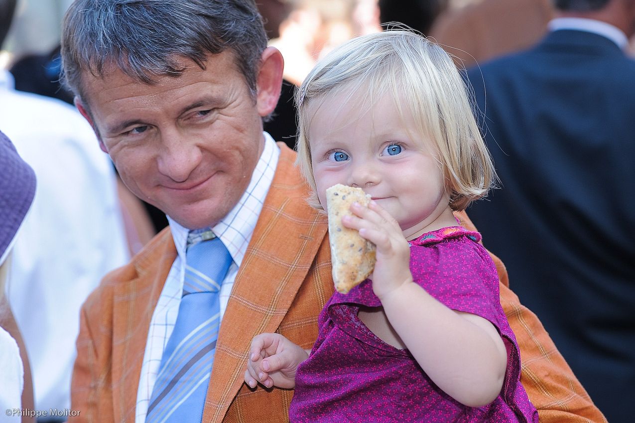 Man in orange blazer holding a blue-eyed toddler girl eating bread at an outdoor event.