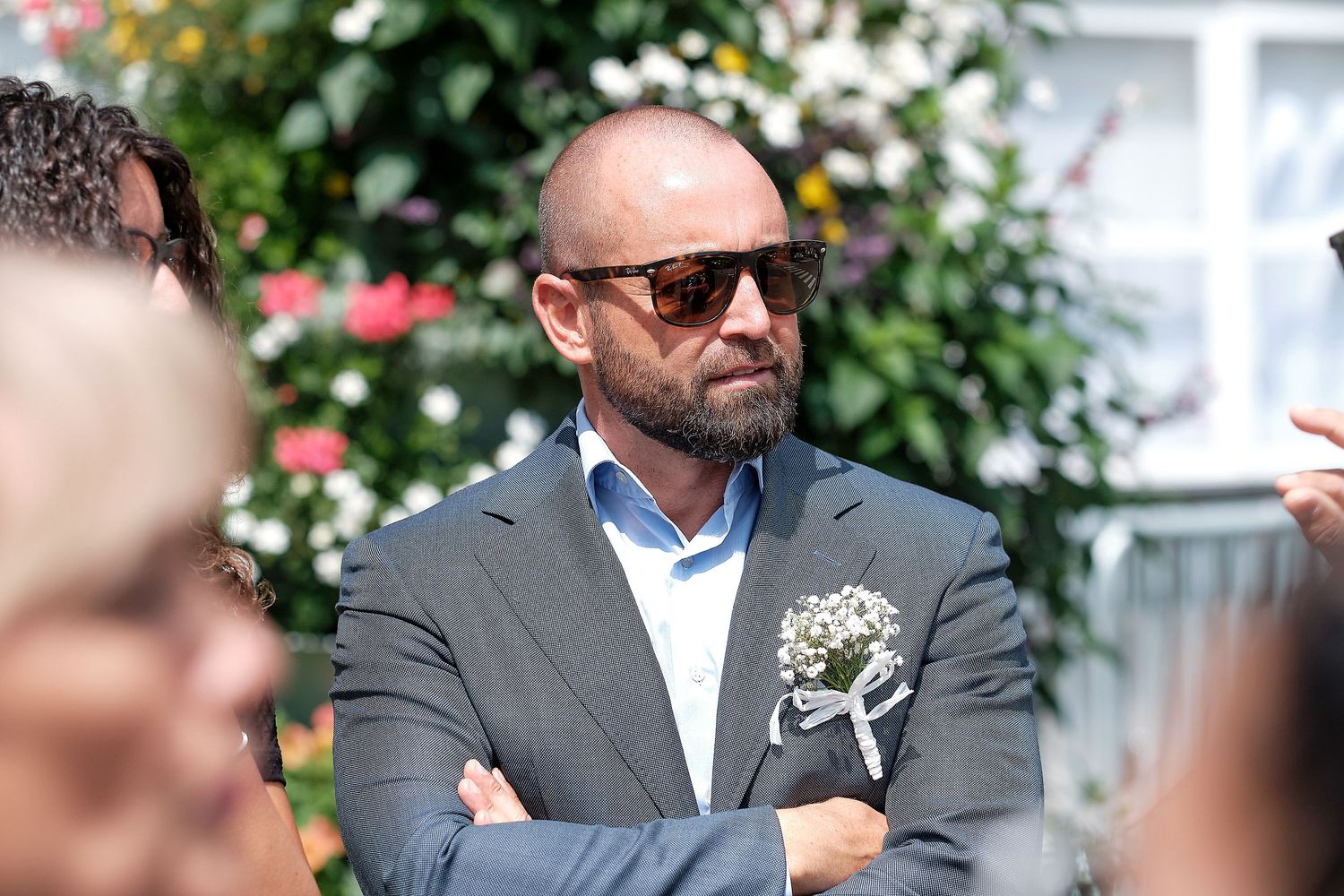 Man in gray suit with sunglasses and boutonniere stands outdoors at a sunny event with arms crossed.