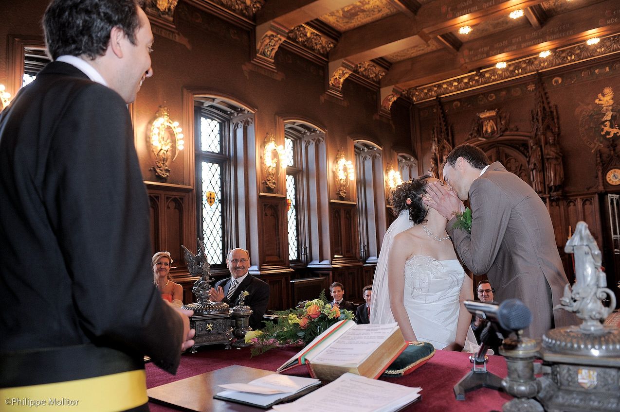 Bride and groom share their first kiss at an elegant wedding ceremony in an ornate wood-paneled hall.