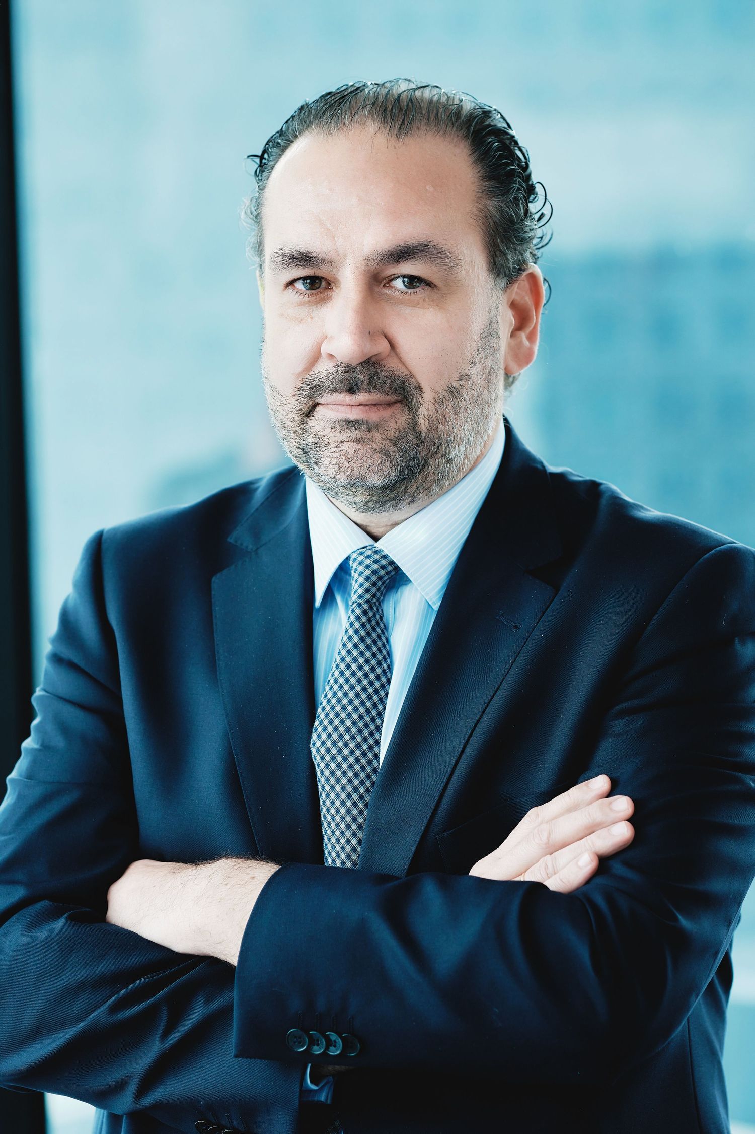 Professional portrait of executive in navy suit with crossed arms against blue background.