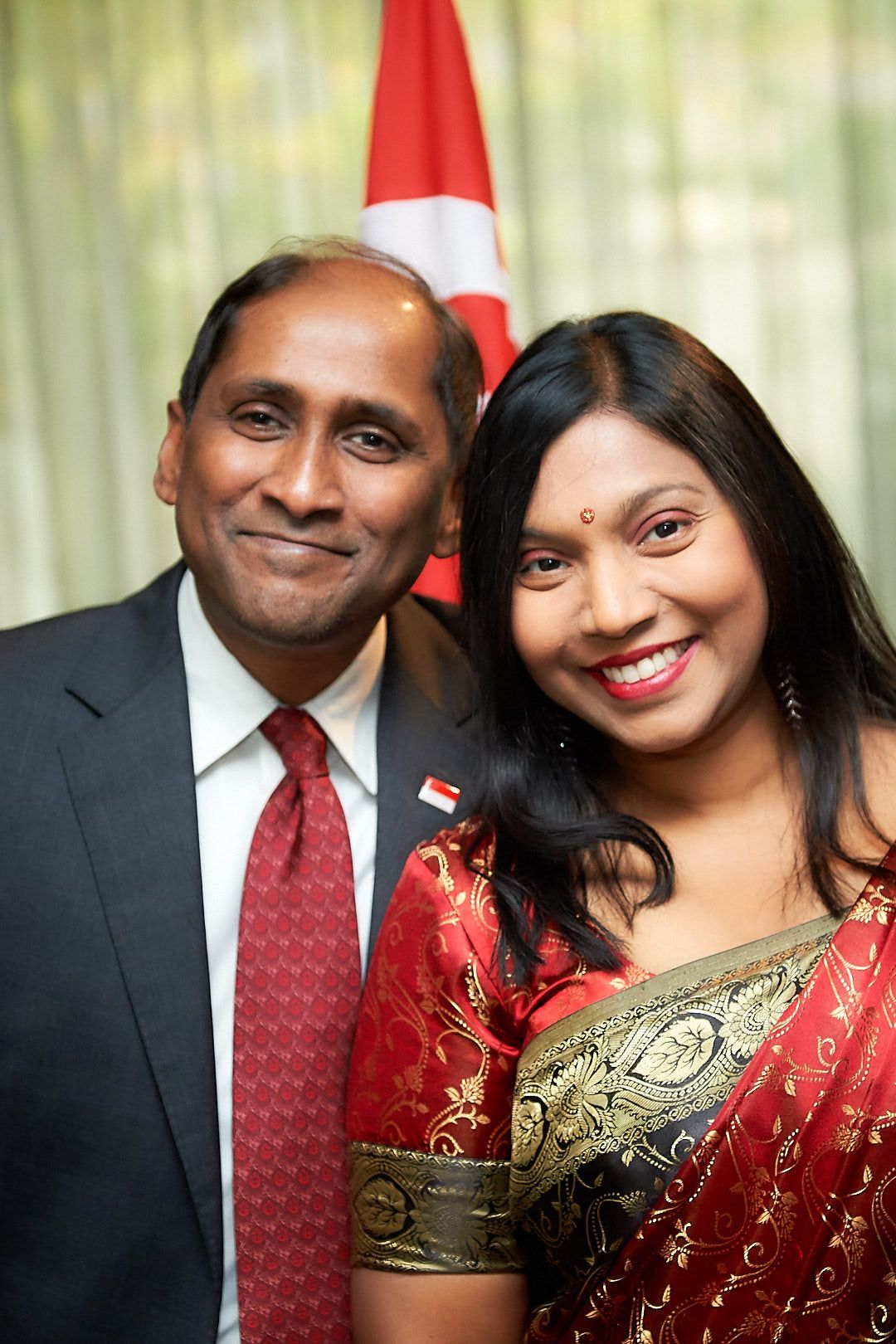 A couple in formal attire poses together against a blurred background with a red flag visible.