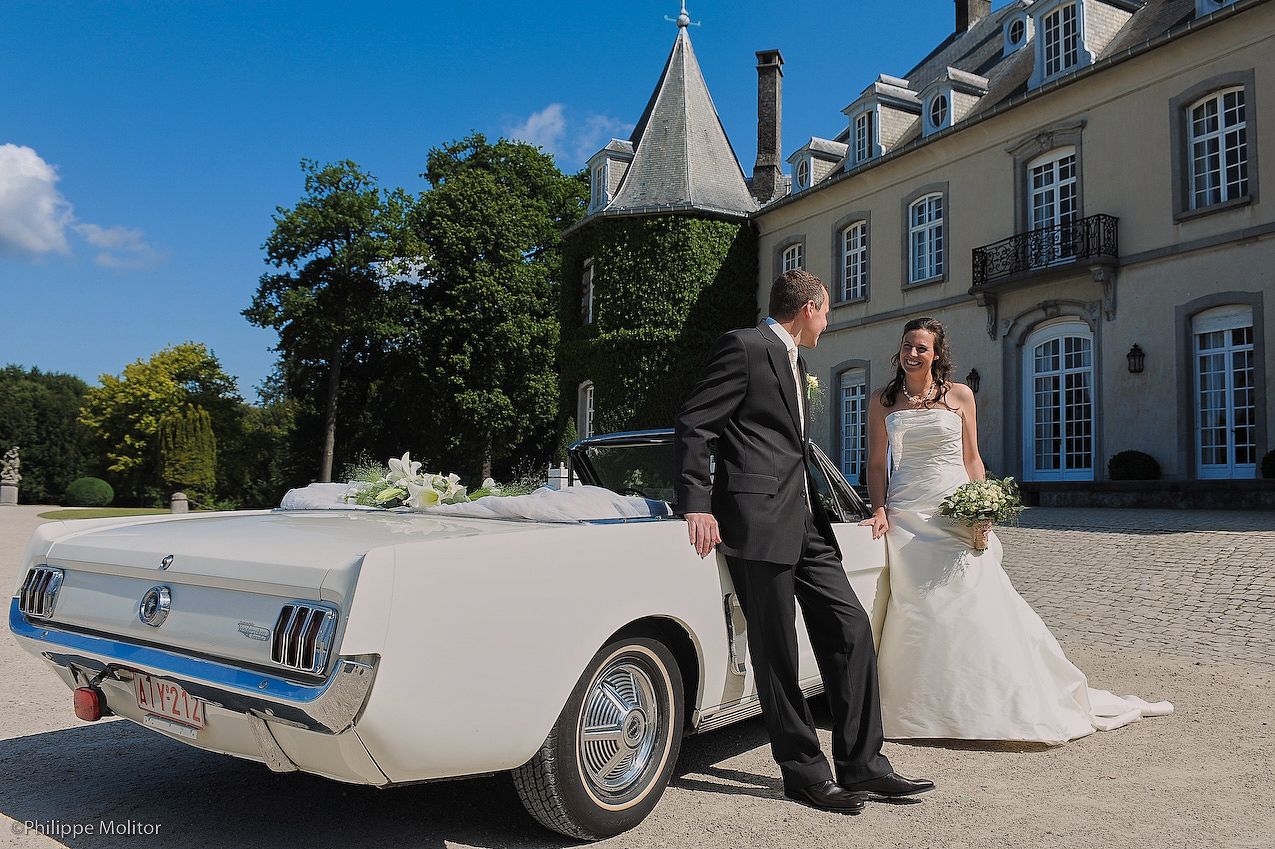 Bride and groom posing beside a classic white Ford Mustang convertible outside an elegant French château on a sunny day.