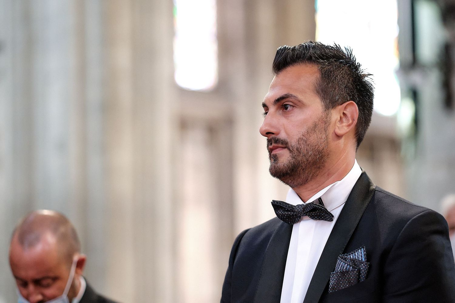 Elegant man in black tuxedo with bow tie and pocket square standing in a grand architectural interior.
