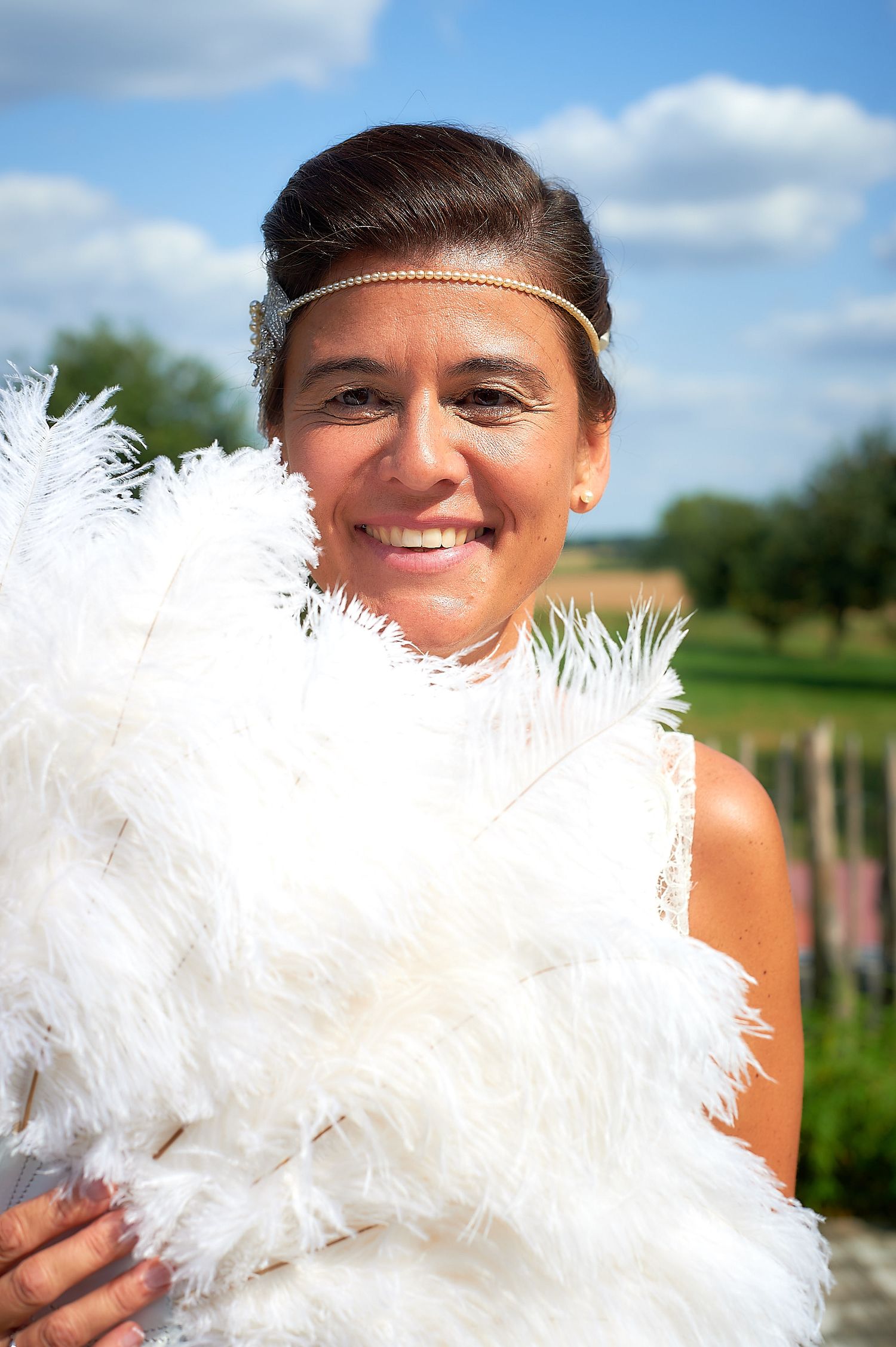 Smiling woman holding white feather boa wearing gold headband outdoors on sunny day.