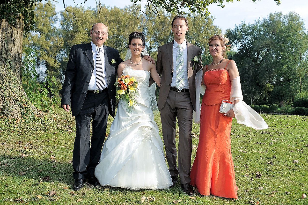 Wedding party of four posing outdoors, bride in white gown with bouquet, groom in gray suit, guests in orange dress.