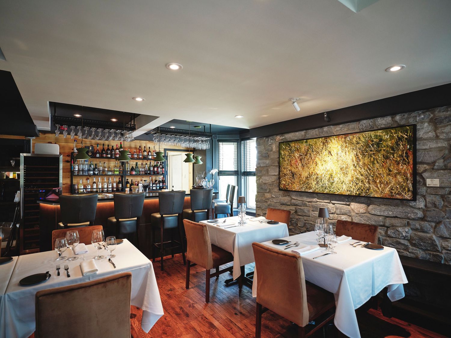 Elegant restaurant interior with white tablecloths, stone wall, large artwork, and a well-stocked bar in the background.