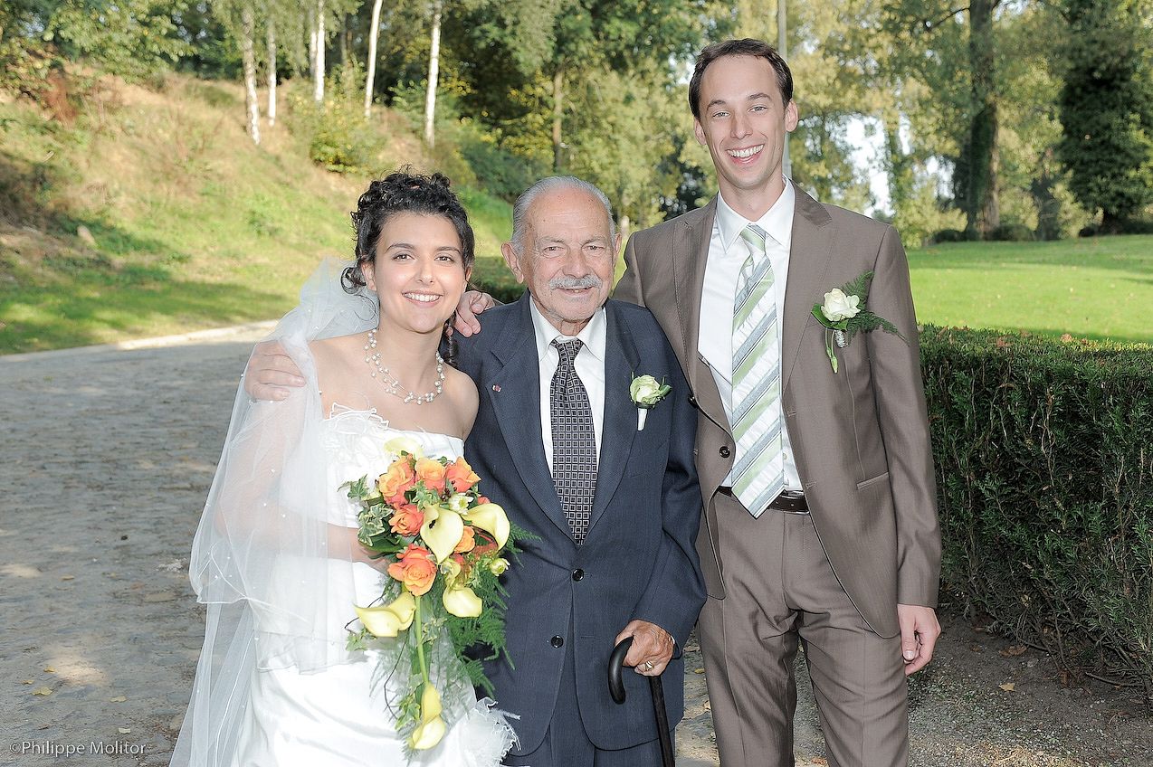 Bride in white dress holding bouquet stands with elderly man and groom in brown suit at outdoor wedding.