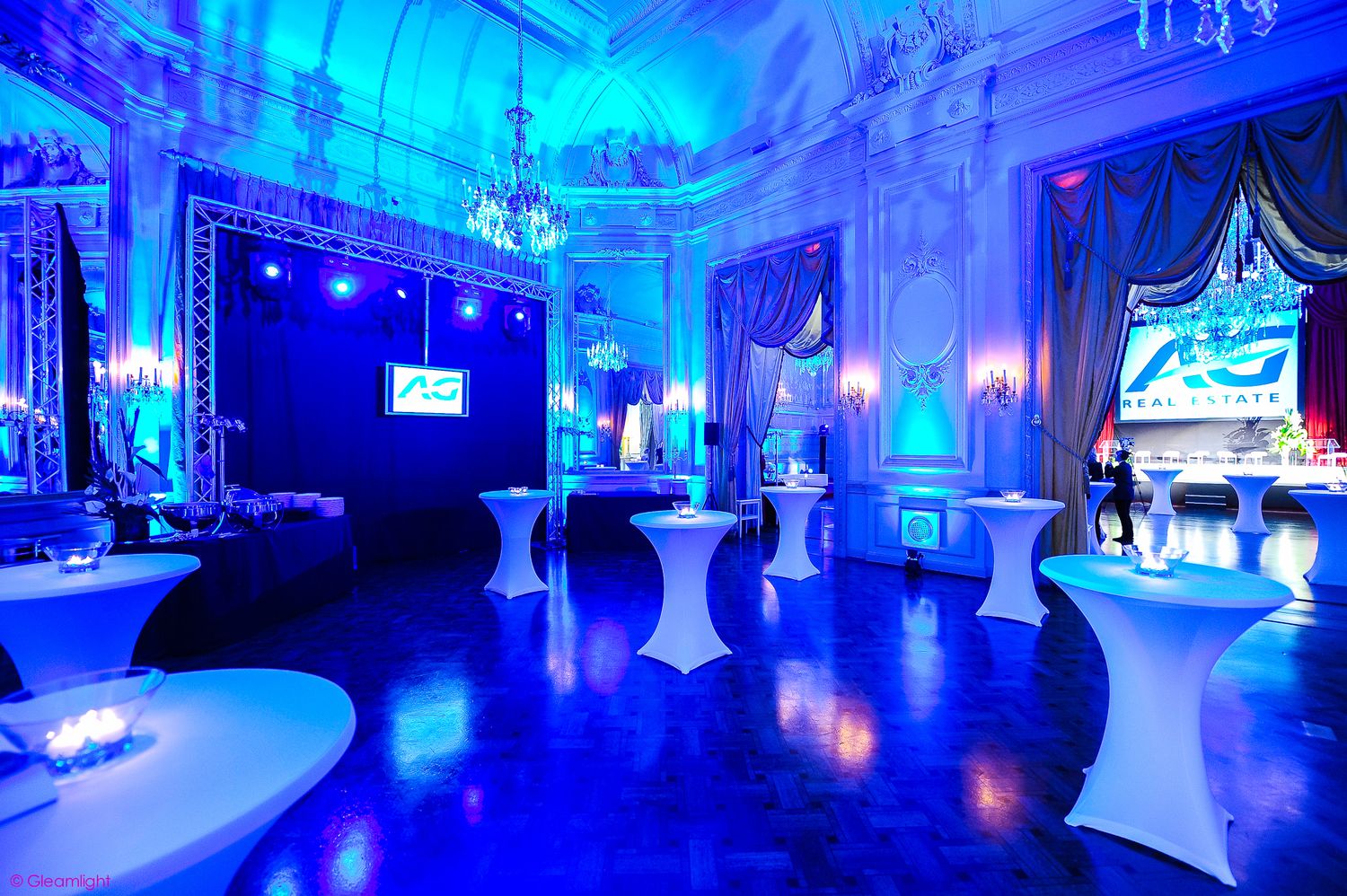 Elegant ballroom venue illuminated in dramatic blue lighting with white-covered chairs and crystal chandeliers.