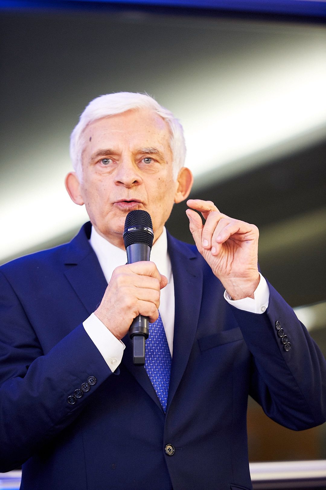 Speaker in a navy suit gesturing while giving a presentation with a microphone.