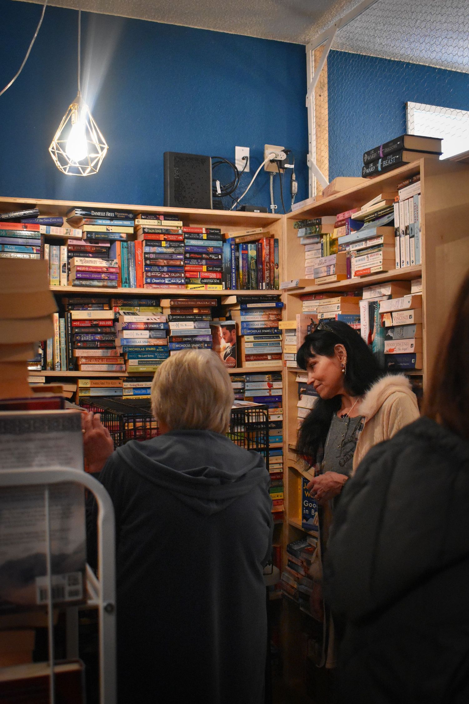 People browse books on wooden shelves inside a cozy bookstore with blue walls and geometric pendant lighting.