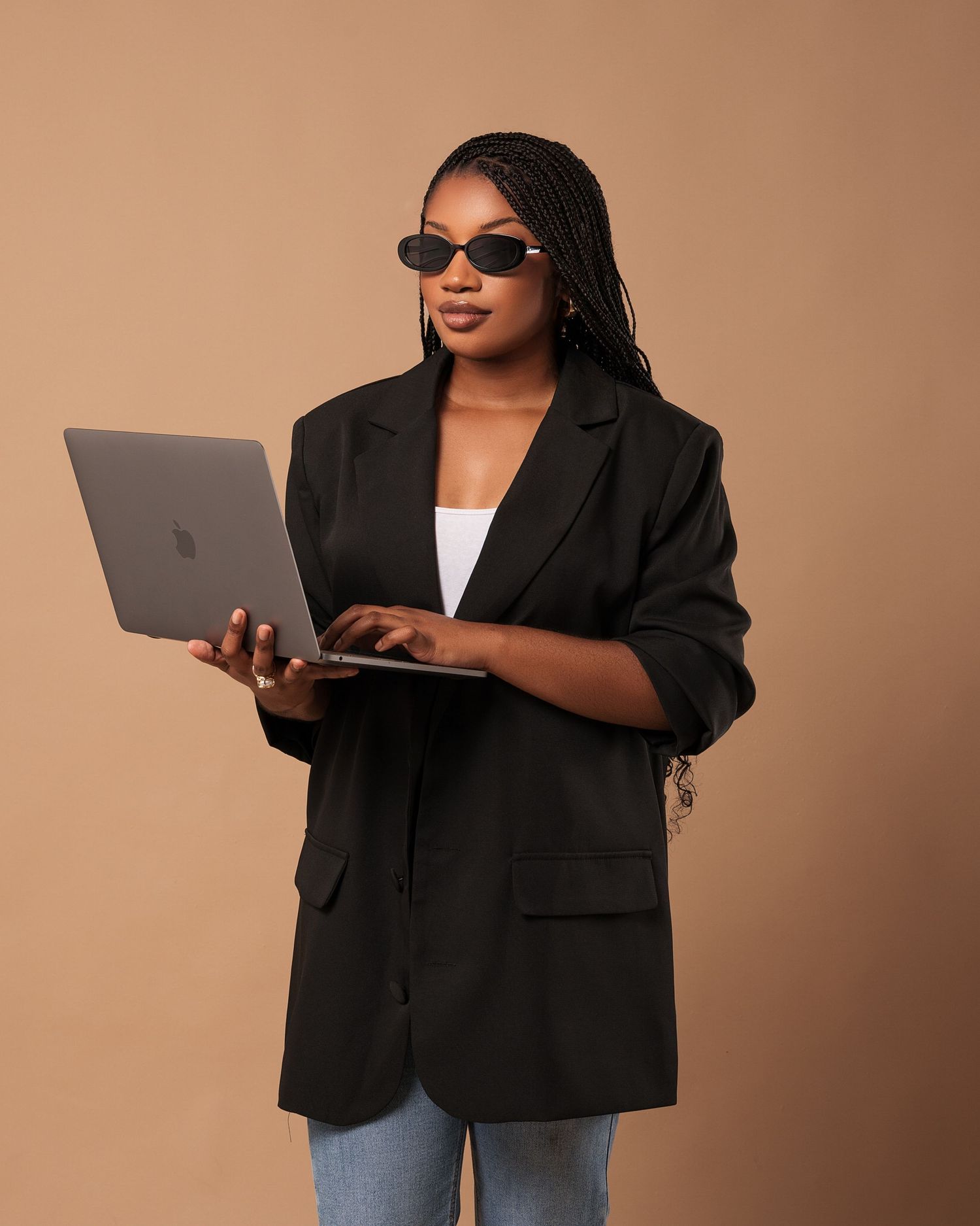 Personne stylée portant un blazer oversize et un jean, posant avec un ordinateur portable sur un fond neutre par MISE VISUALS.