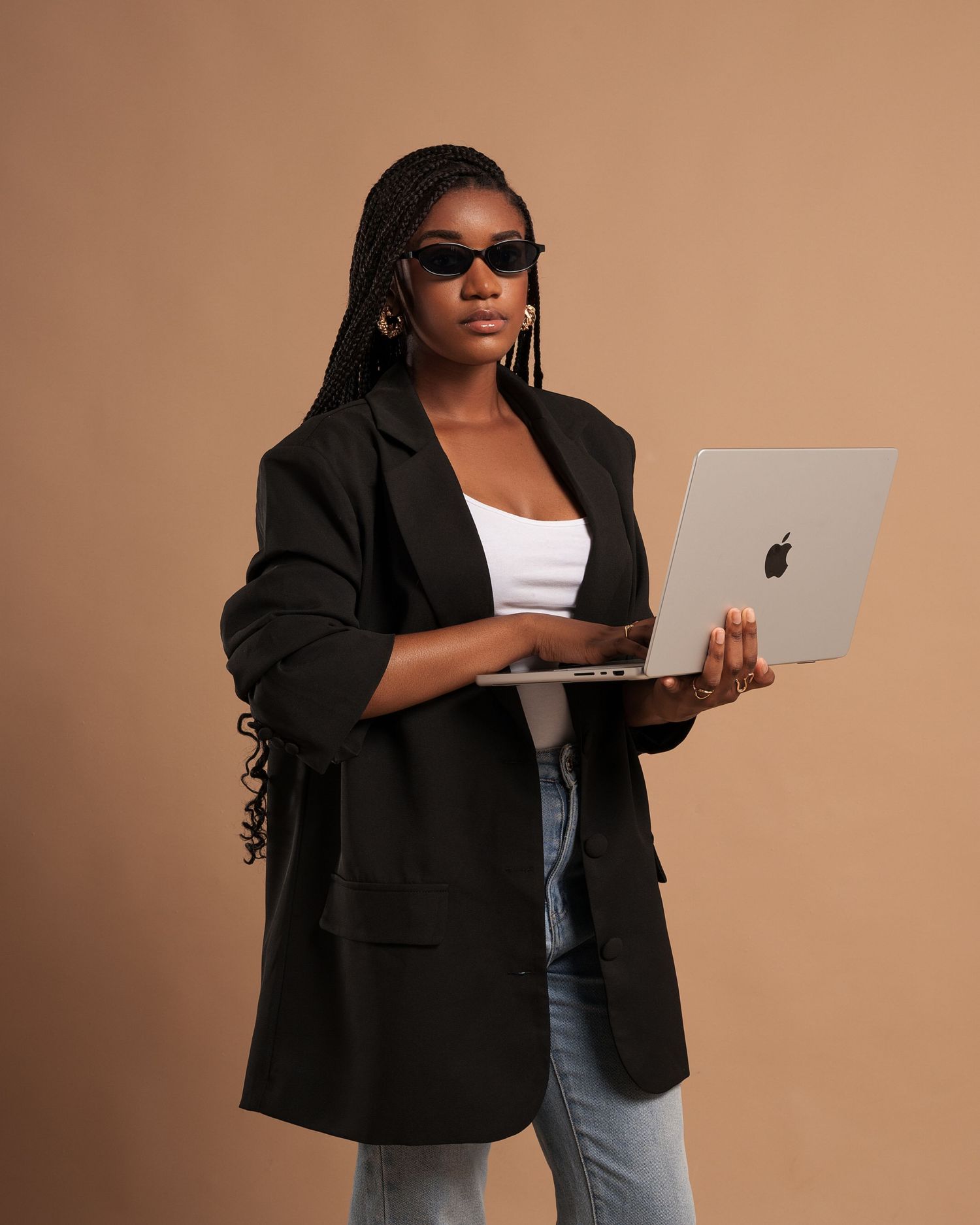 Personne stylée portant un blazer oversize et un jean, posant avec un ordinateur portable sur un fond neutre par MISE VISUALS.