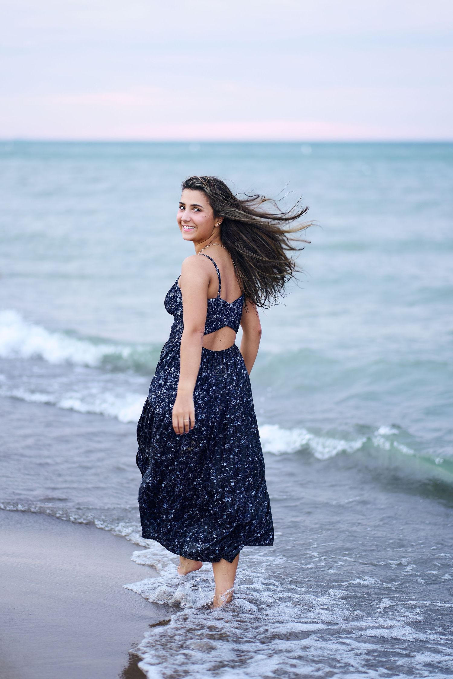 A woman in a navy blue dress walks along the shoreline as waves crash at her feet, her hair flowing in the ocean breeze.