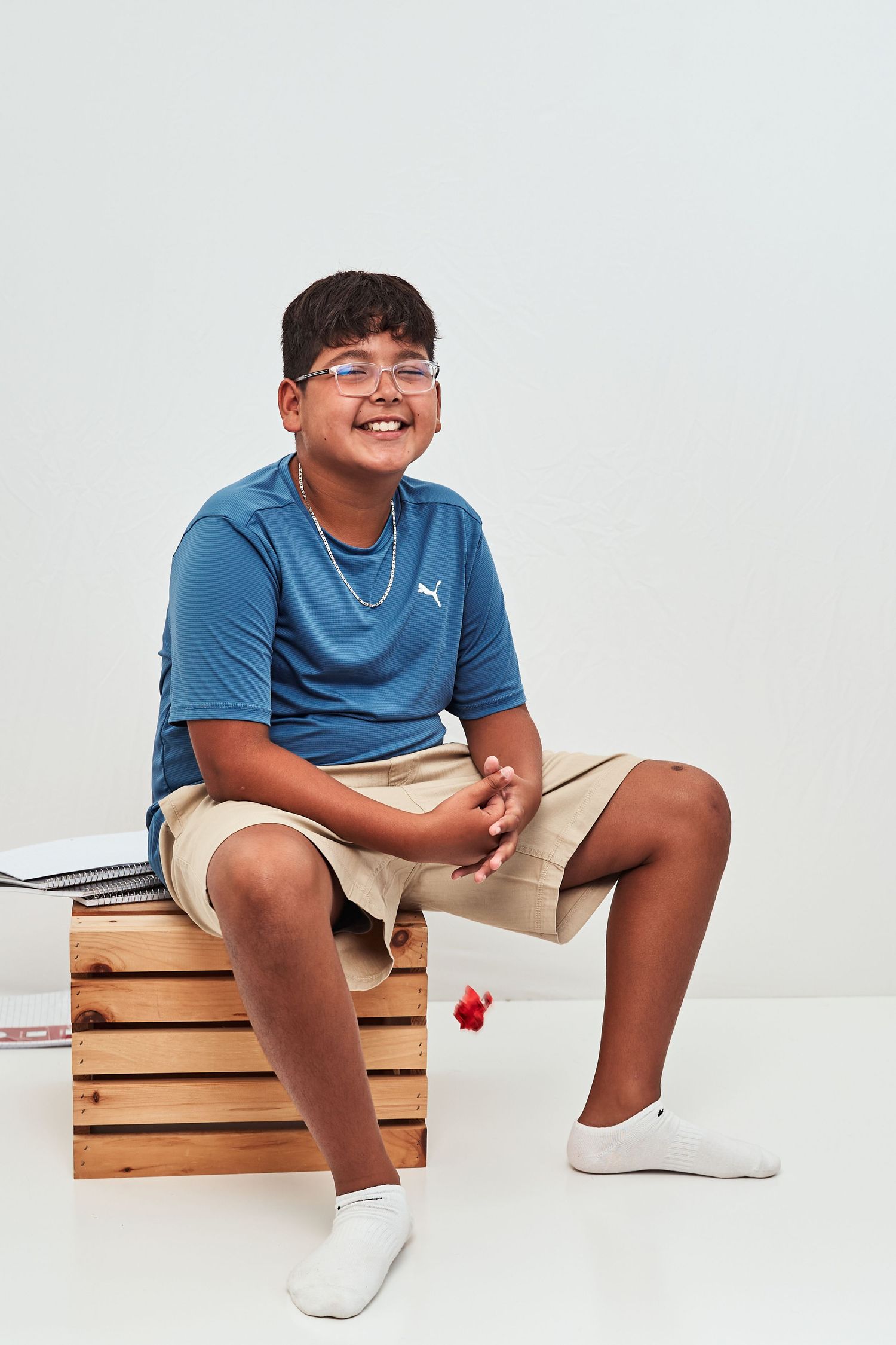 Young person in blue shirt and white sneakers sitting on wooden crates against white background in casual pose.