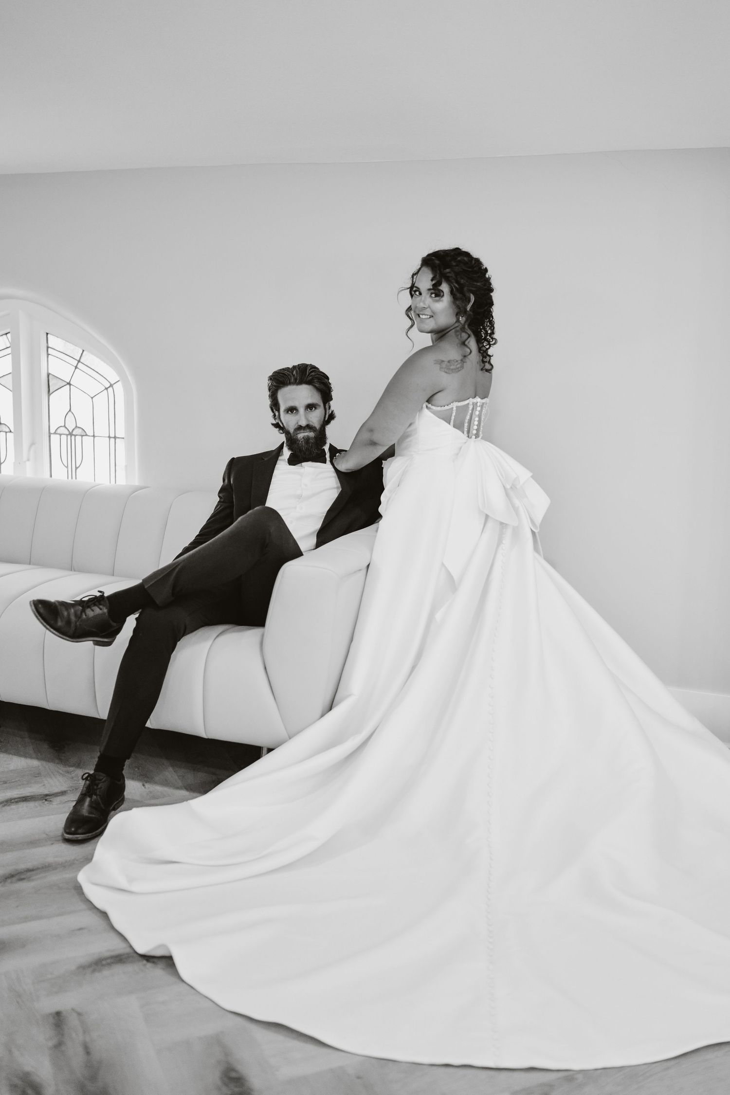 A black and white wedding photo of a couple posing elegantly on a white couch in formal attire.