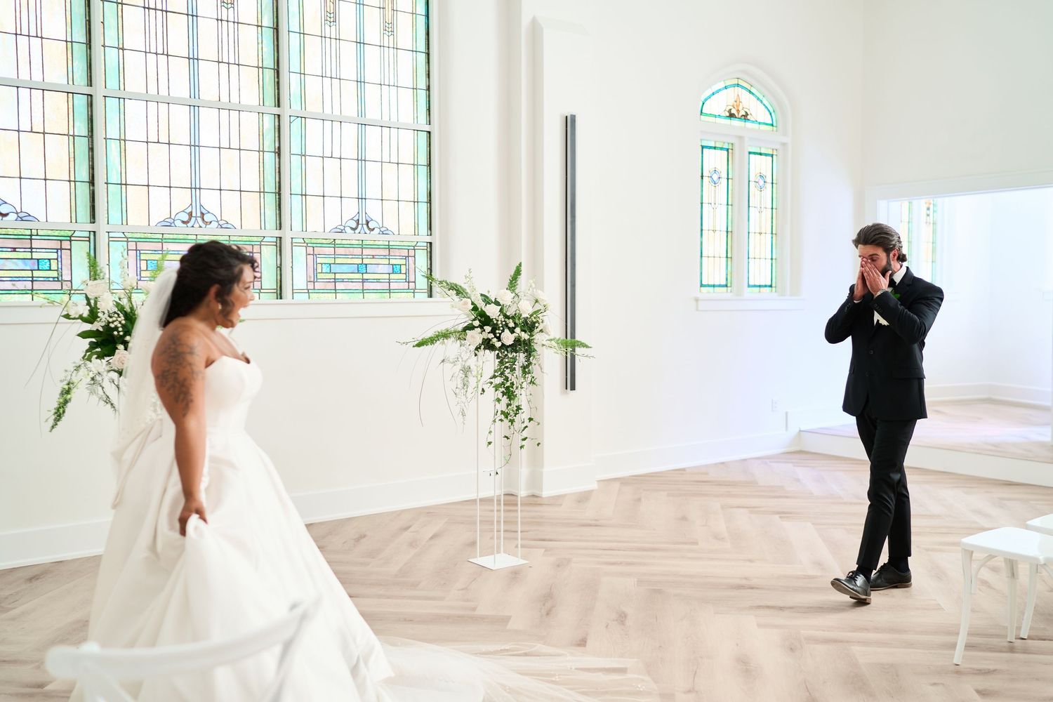 A wedding photographer takes pictures of a bride in a sunlit room with beautiful stained glass windows.