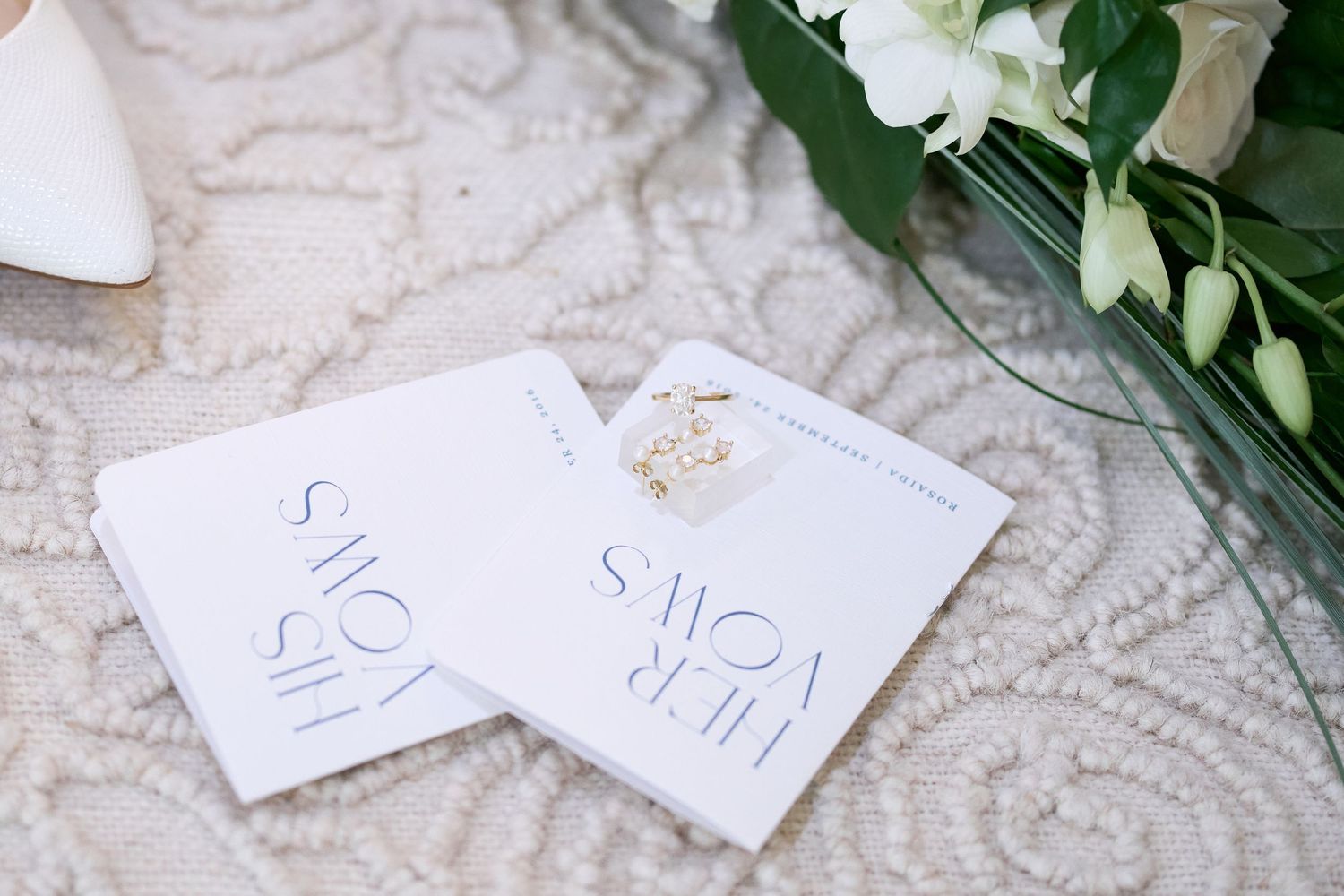 Wedding invitation cards with calligraphy text rest on a textured white fabric next to greenery and flowers.
