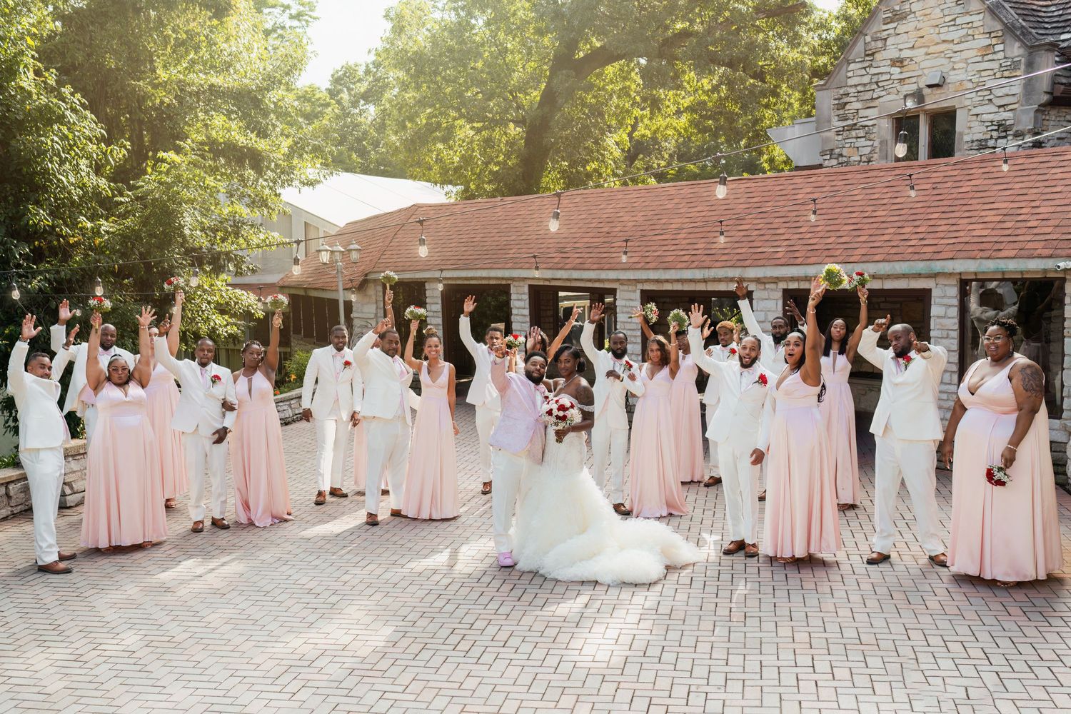 Large bridal party in coordinated pink and white attire poses outdoors at stone venue.