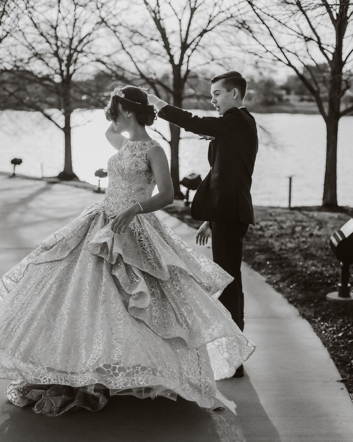 A vintage black and white photo captures a couple on their quinceanera day by a lake with bare trees in winter.