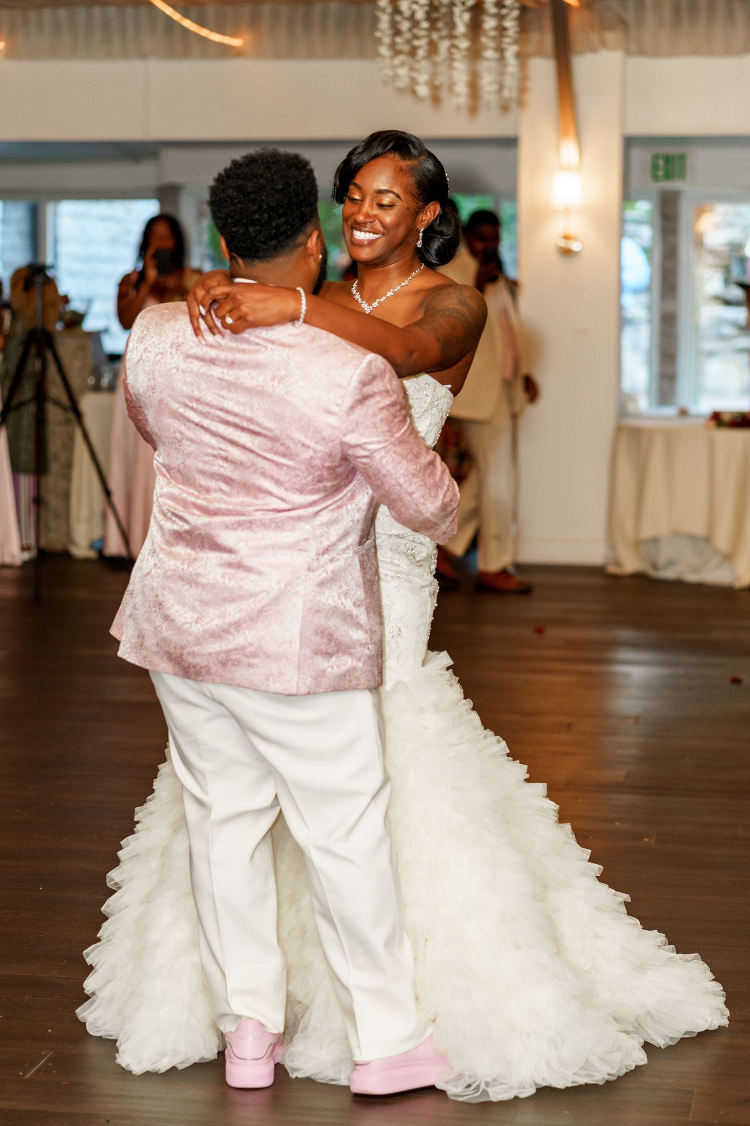 First dance at wedding reception with bride in flowing white gown and groom in pink jacket.