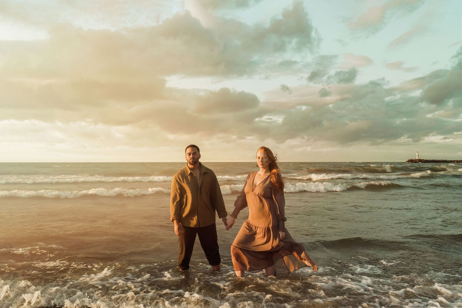 Couple walking hand in hand through ocean waves during sunset on beach.