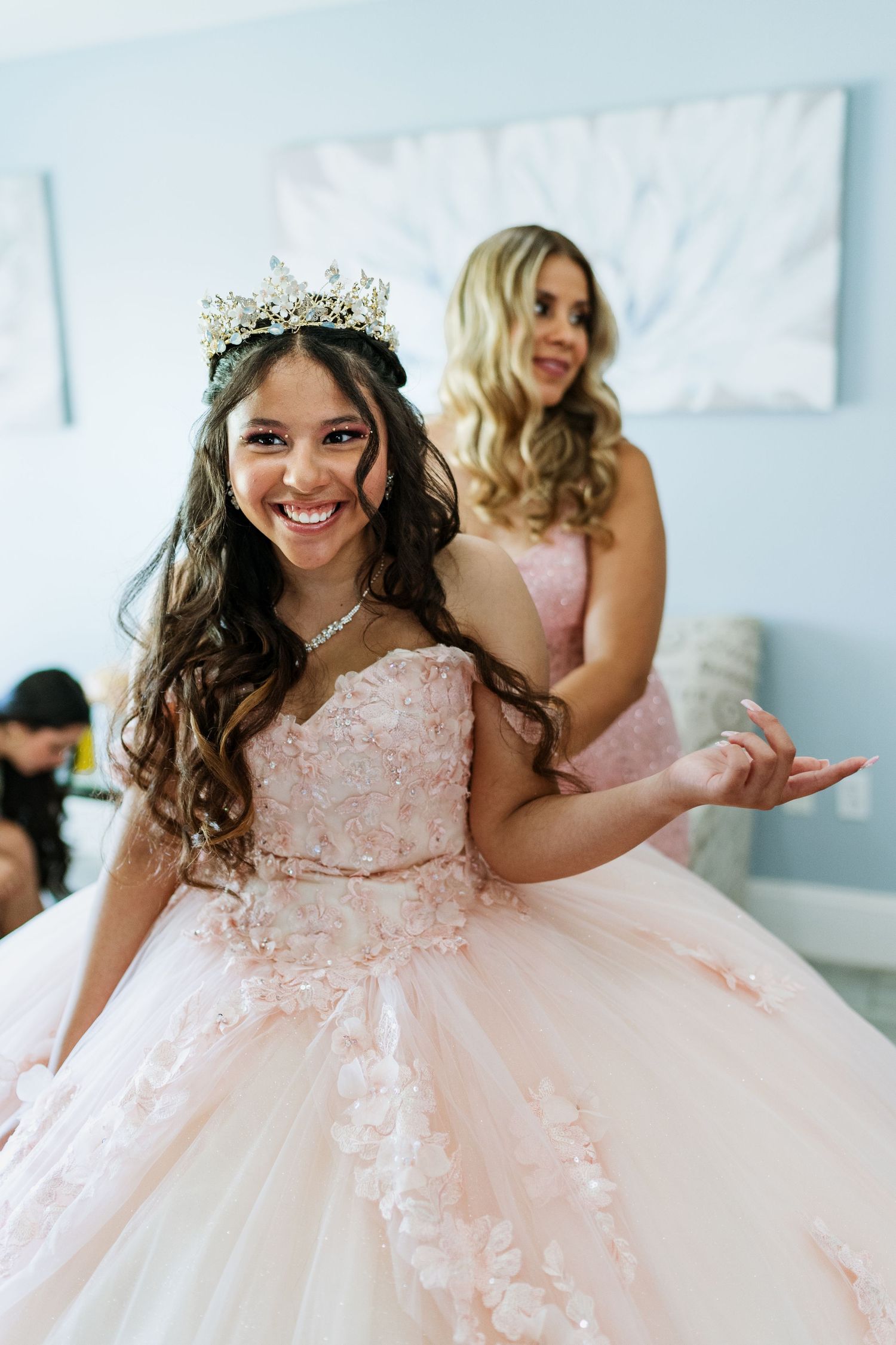 A person in a blush pink ballgown and crystal crown smiles joyfully while getting ready.