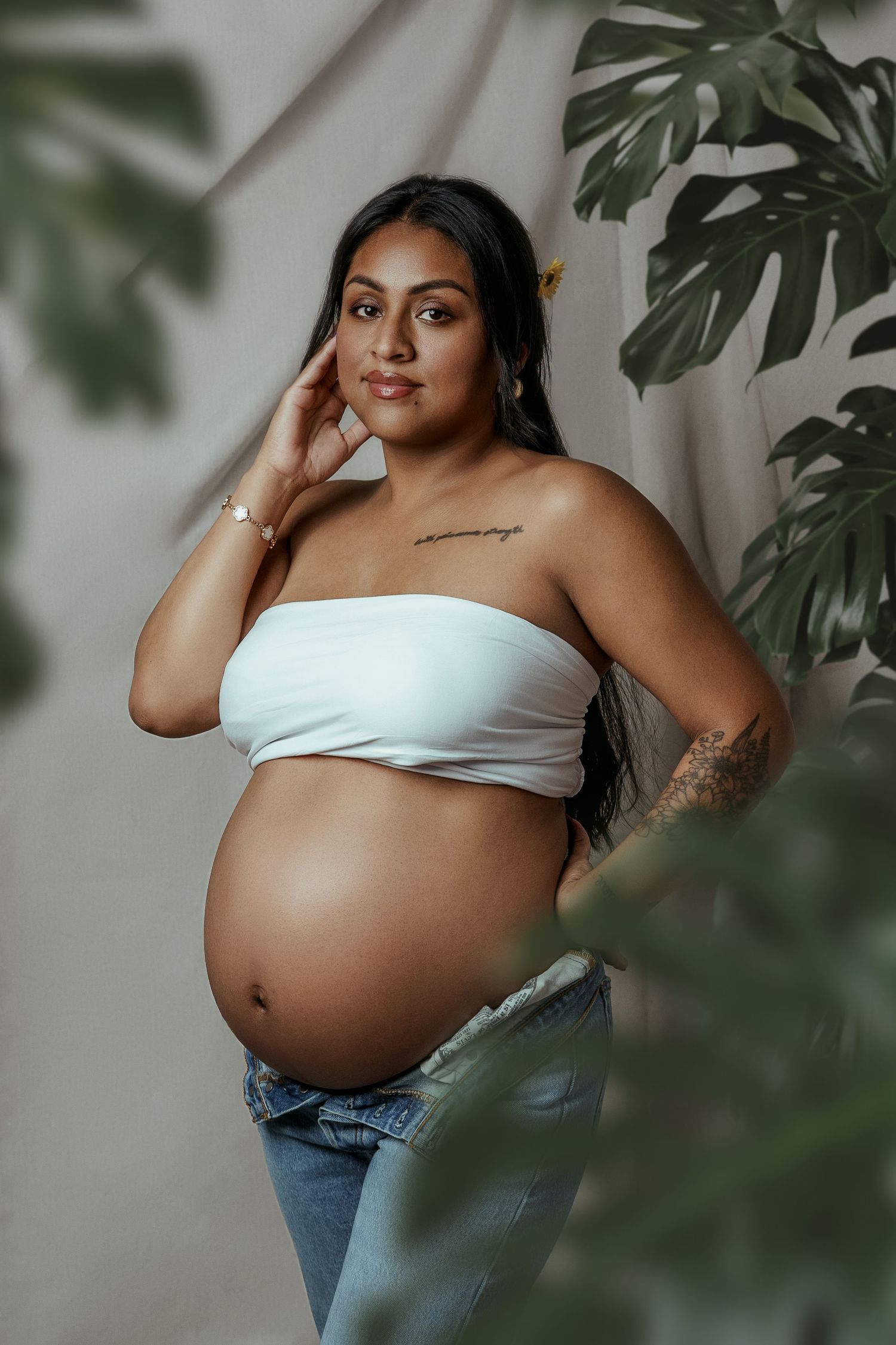 A series of maternity photos showing a pregnant person in white crop top and jeans posing with monstera leaves.