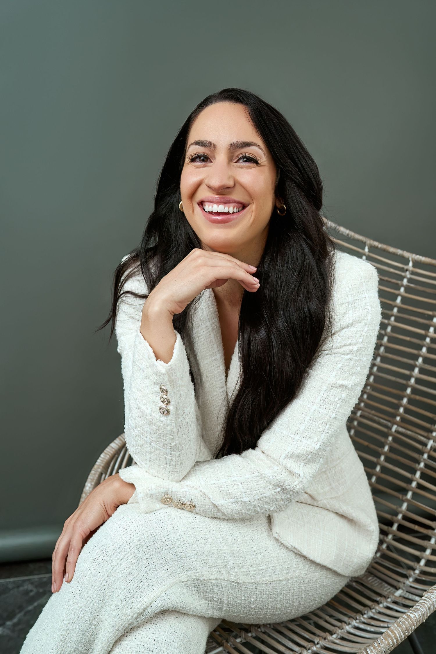 A person in a cozy white sweater lounges on a wire chair while smiling warmly against a dark gray background.